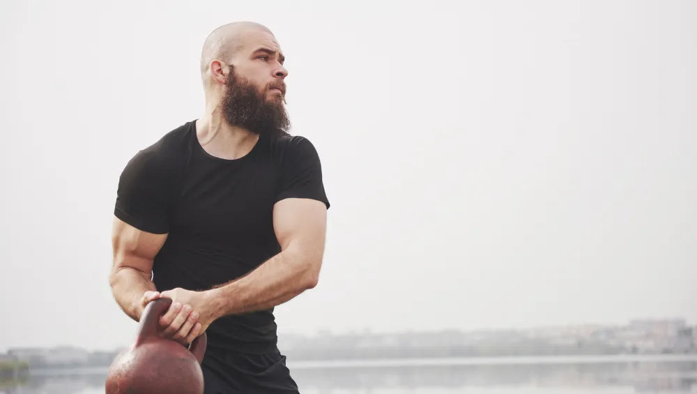 Homme sec et musclé avec un t-shirt et short noir, boule à zéro ainsi qu'une barbe mi- longue attrapant à 2 mains un kettlebell et s'exerçant avec