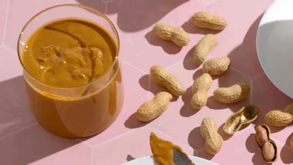 Un verre contenant du beurre de cacahuète avec à coté des cacahuètes en moceaux sur une table rose 