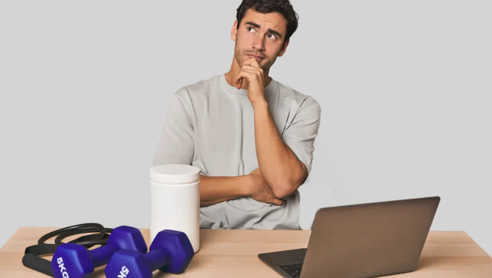 Homme assis en t-shirt gris en train de réfléchir devant un bureau ou est posé un ordinateur portable, des haltères, une corde ainsi qu'un pot blanc avec en arrière plan un fond gris