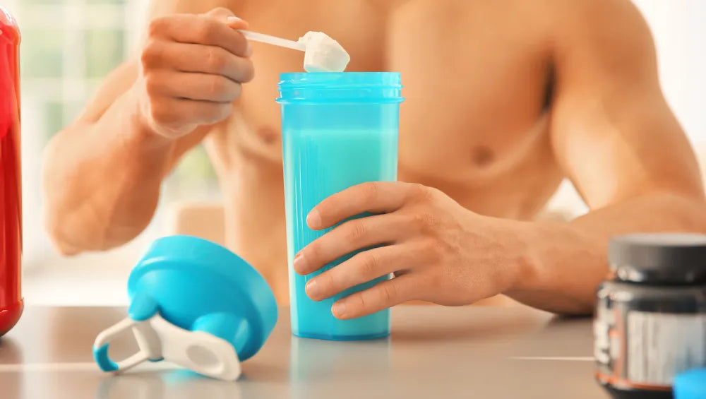 Homme musclé torse nu consommant des BCAA/EAA sur une table, il met les acides aminé dans un shaker bleu ciel 