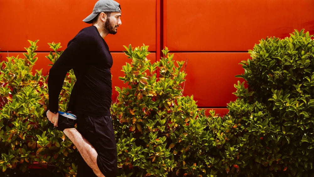 Homme en t-shirt manche longue, short noir et casquette grise en train de s'échauffer en vue d'un footing sur un fond rouge et des buissons en arrière plan