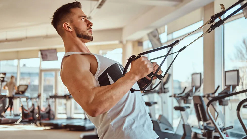 Homme dans une salle de sport ensoleillé en train d'éffectuer un exercice de tirage pour le dos avec un TRX