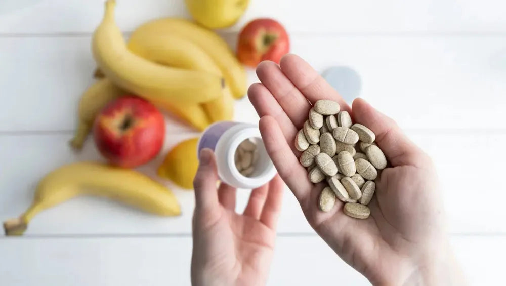 Une main contenant plusieurs comprimé (ZMA) avec en fond sur une table blanche plusieurs bananes et pommes