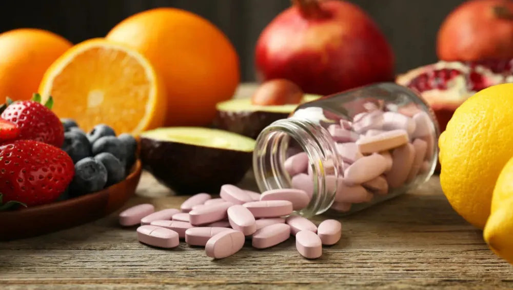 pilulier en verre renversé faisant tomber plusieurs pilule de couleur rose sur une table en bois garnis tout autour de plusieurs oranges, grenades et fruits rouge