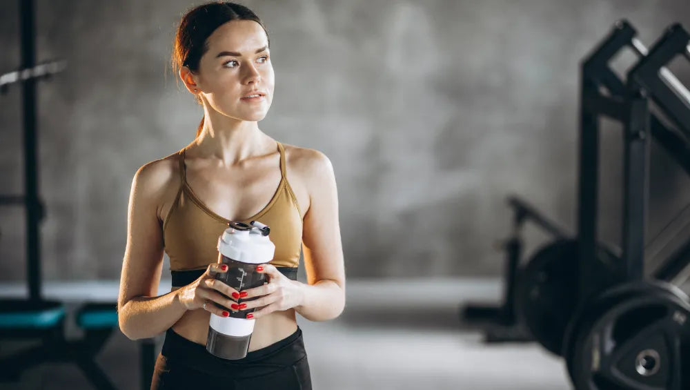 Une femme en crop top marron dans une salle de sport avec un fond gris portant un shaker et regardant au loin, légèrement de profil