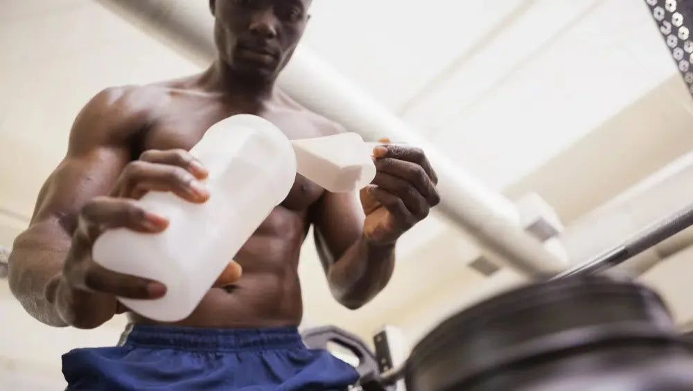 Homme musclé torse nu en train de mettre une scoop de protéine dans son shaker dans une salle de sport