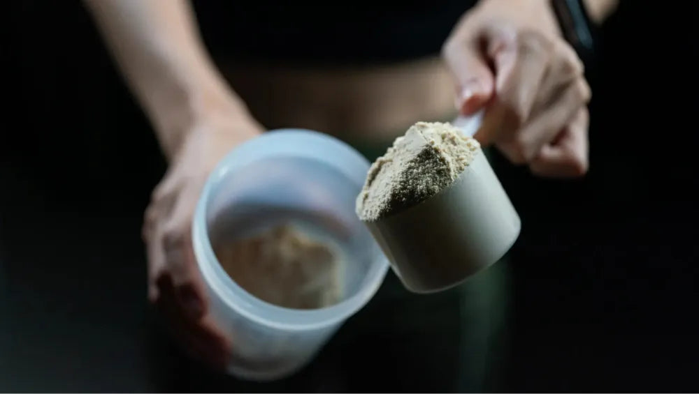 Une femme sur un fond noir en train de montrer une scoop remplis de protéine avec en fond un shaker contenant également de la protéine 