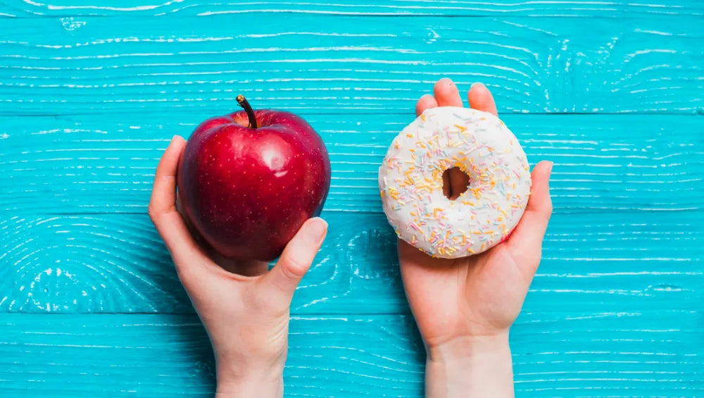 Une pomme dans une main gauche et un donut dans une main droite sur un fond bleu ciel en bois