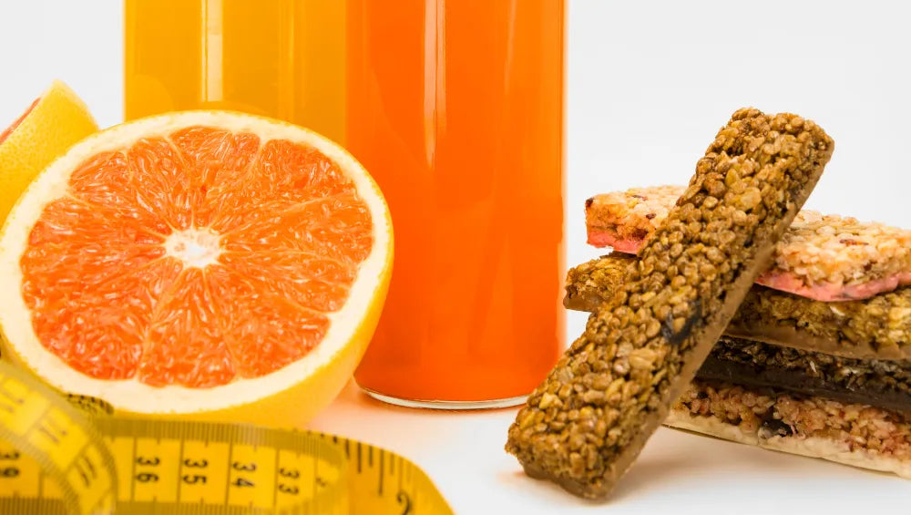 Orange coupé en 2 avec une bouteille en verre en arrière plan de jus d'orange, quelques barre de céréales ainsi qu'un mètre souple