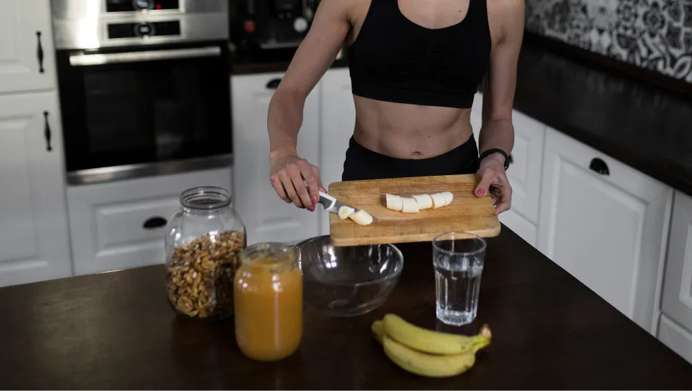 Femme dans une cuisine en train de mettre des bananes coupée sur une planche à découpé dans un grand saladier, sur la table un pot de beurre de cacahuète avec des noix à coté