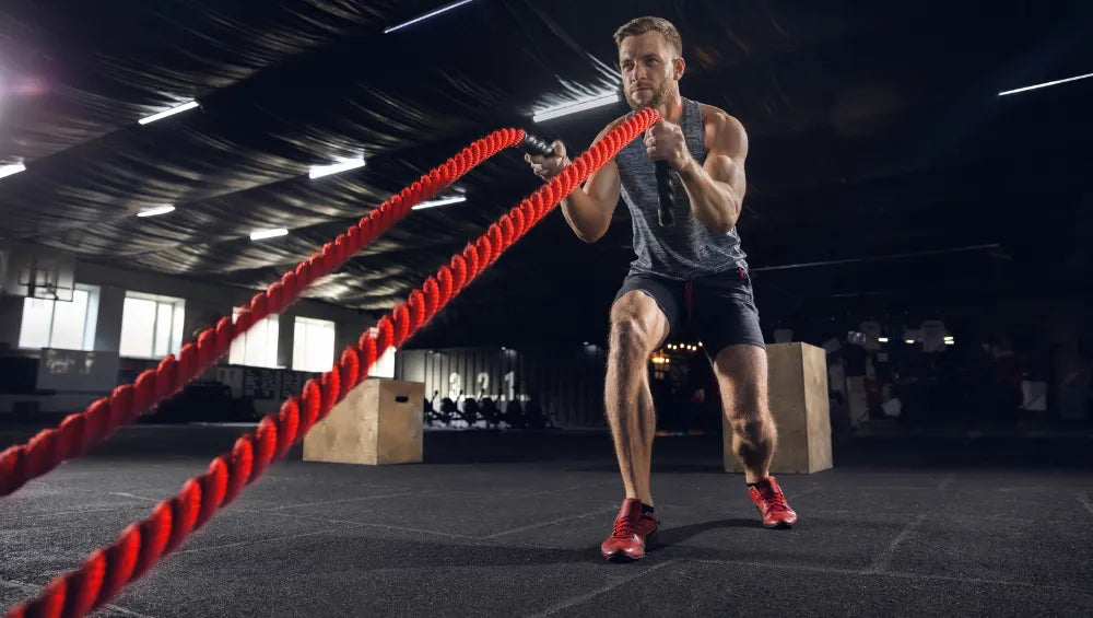 Homme dans une salle de sport coté crossfit streetworkout, en train de faire un exercice d'agitation de corde avec plusieurs step derrière lui