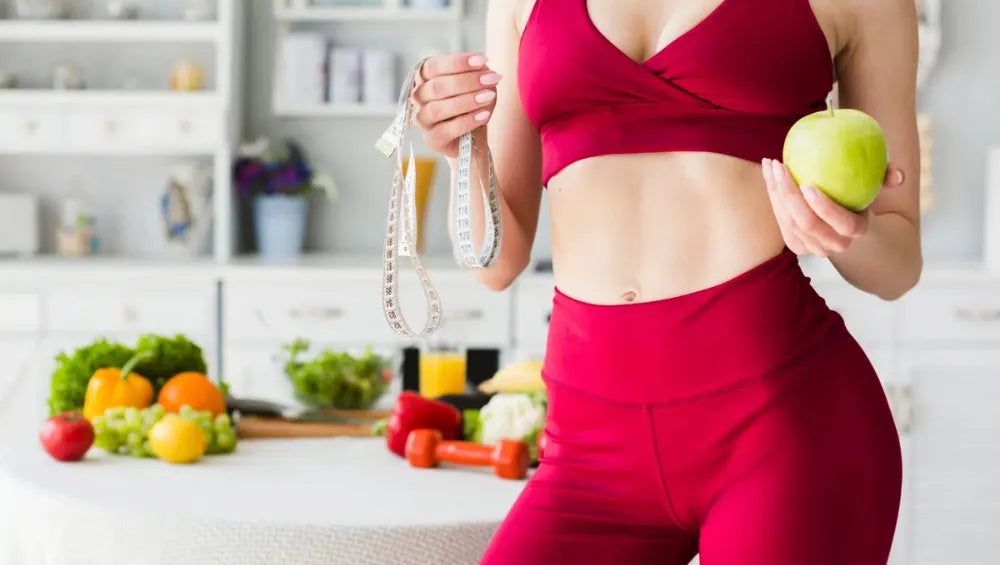 Femme habillé en legging et crop top rouge dans une cuisine avec sur la table en arrière plan plusieurs fruits et légumes tenant dans une main une pomme et dans l'autre un mètre souple