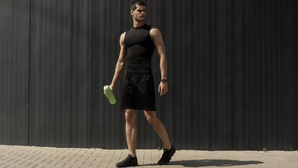 Homme en débardeur et short noir ce tenant droit légèrement de profile avec une gourde verte dans sa main droite dehors sur un fond noir pendant une journée ensoleillé