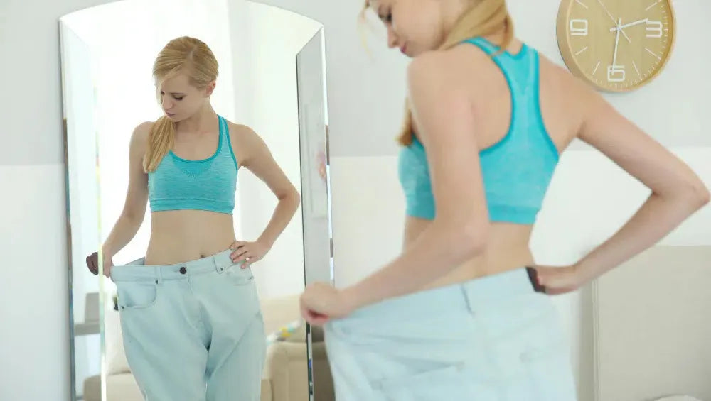 Femme en crop top bleu et jean "flottant" devant le miroir de chez elle regardant la partie du jean qui ne lui vas pas 