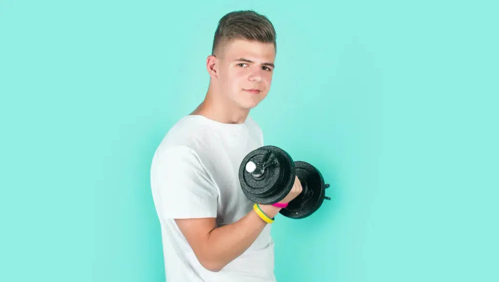 Jeune adolescent debout légèrement de profil en t-shirt blanc avec un haltère dans la main en train d'exécuter un mouvement de curl biceps sur un fond bleu ciel